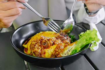 Close-up shot of hands using cutlery to enjoy a portion of Chiffon Egg Bulgogi (Omurice) at a restaurant table. Korean food and lifestyle
