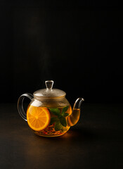 Steaming glass teapot with orange slices, herbs, and mint on dark background. Warming herbal tea for winter wellness.