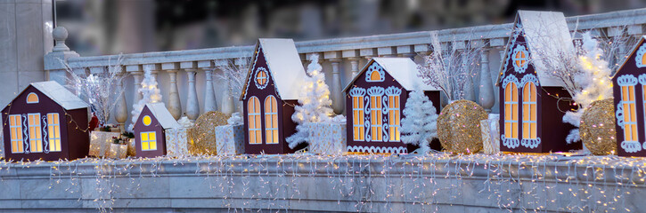 Festive Christmas village display with illuminated houses, white tree, gift boxes, and sparkling lights, creating cozy holiday atmosphere. Banner