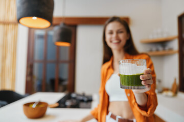 Smiling woman holding a green smoothie in a stylish kitchen, showcasing healthy eating and a vibrant lifestyle in a bright, modern setting