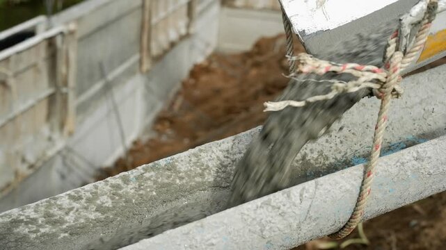 A dynamic, close-up shot captures the process of pouring fresh, wet concrete into a makeshift formwork channel on a construction site. The thick, viscous mixture is rapidly flowing, creating a motion 