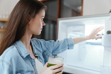 Young woman in casual denim shirt reaching for healthy green smoothie in refrigerated kitchen storage, promoting healthy lifestyle and nutrition