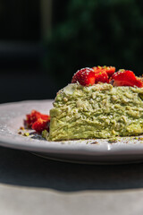A piece of Napoleon cake with pistachio, decorated with strawberries on a table in a bowl in the sun in a restaurant. Close-up photo