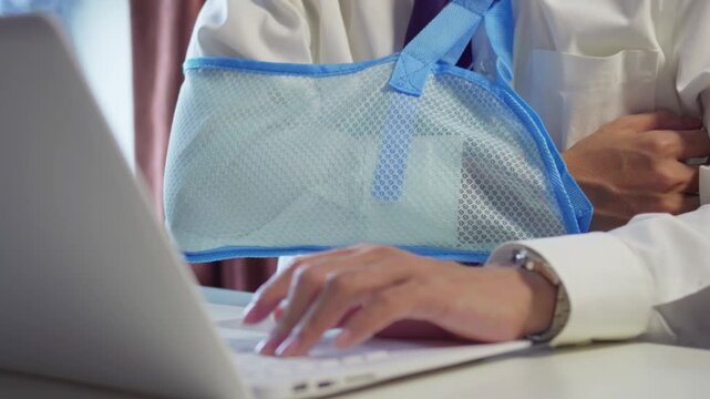 Close up of injured Asian businessman wearing hand splint while working in modern office, typing on laptop with bandaged wrist. Workplace injury, rehabilitation, recovery, and healthcare concept.