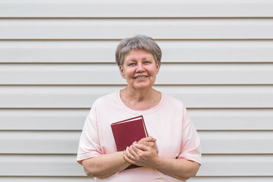 Smiling elderly woman with short gray hair holding a red book while standing against light siding wall, wearing pink T-shirt, looking friendly and positive, concept of lifelong learning and happiness