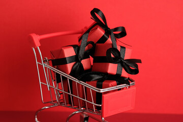 Mini shopping cart filled with red gift boxes tied with black ribbons on red background. Black...