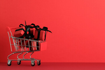 Mini shopping cart filled with red gift boxes tied with black ribbon on red background. Festive...