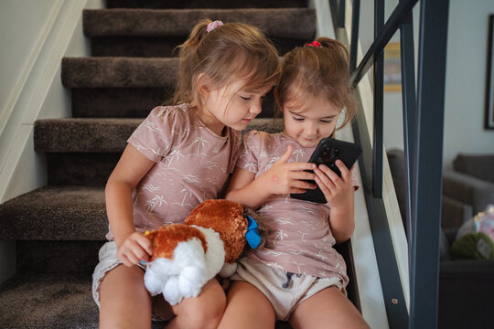 Two preschool age little sisters playing with smartphone sitting on stairs at home