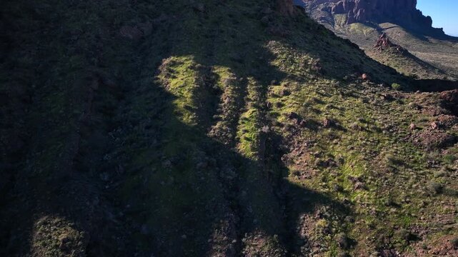 Aerial view of Superstition Mountain's rugged terrain, with green vegetation clinging to the steep slopes under a clear blue sky, Apache Junction, Arizona, United States.