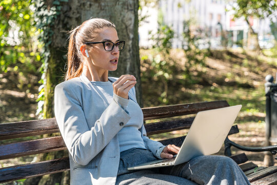 Young businesswoman on a park bench, video call on her laptop, remote work with wireless earbuds freelance online meeting flexible telework digital-nomad productivity and positive client communication