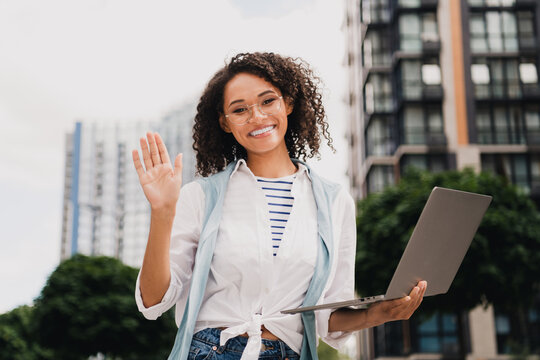 Smiling woman with laptop waves at camera in sunny urban city scene showcasing casual fashion and modern work vibe outside stylish buildings