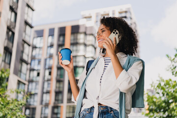 Fototapeta premium Young woman in urban city scene talking on phone with coffee cup stylish casual fashion and modern lifestyle out in the sun