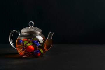 Glass teapot with strawberry, mint leaves, and blue cornflower petals on a dark background. Herbal fruit tea rich in antioxidants and vitamins.