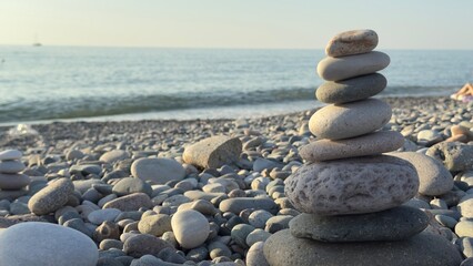 Balanced stack of smooth stones on a pebble beach with ocean waves and sailboat in the background under a clear sky at sunset