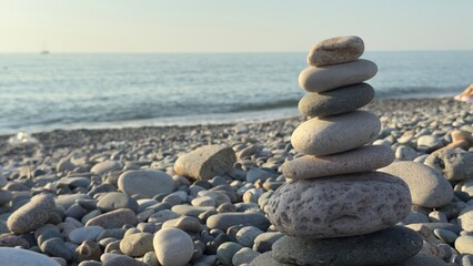 Balanced stack of smooth stones on a pebble beach with ocean waves and sailboat in the background under a clear sky at sunset