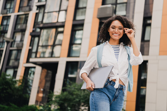 Young woman student with laptop walking outside modern city building smiling and ready for study and travel in casual fashion
