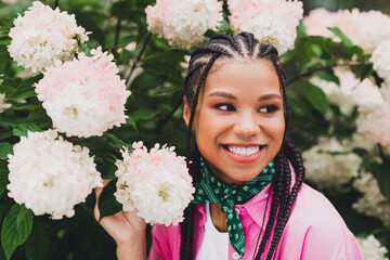 Portrait of a young woman with braids in a pink jacket among hydrangeas smiling in a sunny garden...