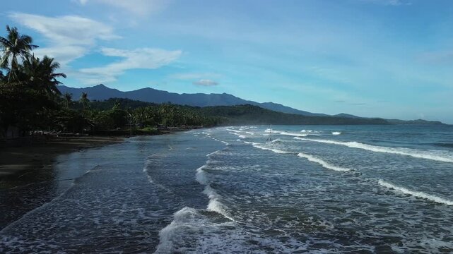 Low aerial flight over restless sea with surging waves to dark sandy shore lined with palms, boats and houses, backed by mountains in Aporawan, Little Siargao ng Puerto, Palawan, Philippines.