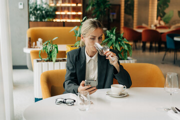 Attractive blond business woman in a modern restaurant drinking water and using her mobile phone.