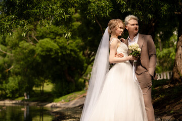 Couple in wedding attire posing by the water under trees in a park