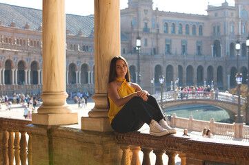 A young, dark-haired, attractive Latin woman wearing a yellow shirt and black trousers is sitting...