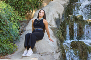 A young, pretty Spanish woman with dark hair sitting on the rocks next to the waterfall in the park. The woman is wearing black jeans and a black T-shirt with a scarf around her neck.