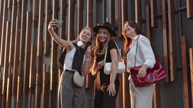 Three stylish women standing near wooden wall. Smiling while taking selfie. Female in white top holding smartphone and making photo. People enjoying cheerful moment and posing to camera.