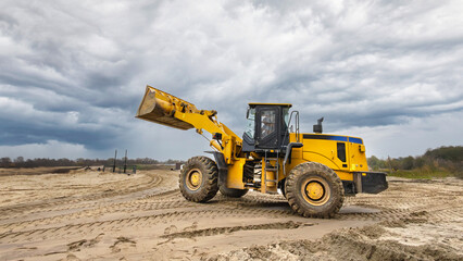 Obraz premium A yellow loader operates on sandy terrain under a gray sky, preparing the site for upcoming construction activities near the water