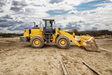 A yellow front-end loader is working on a construction site, moving dirt while dark clouds loom overhead, indicating changing weather conditions