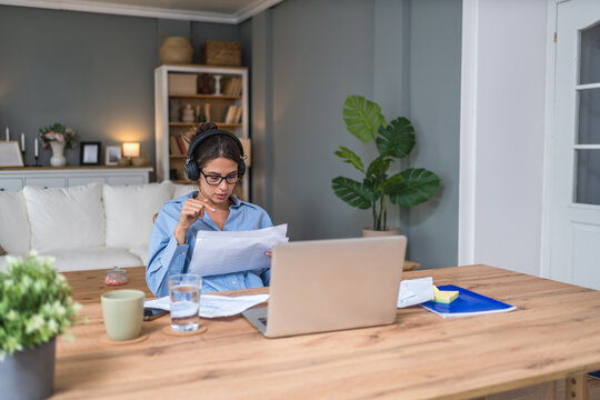 Focused freelance businesswoman in a home office wearing headphones, reviewing financial reports while attending an online session on her laptop budget planning, productivity and efficient remote work