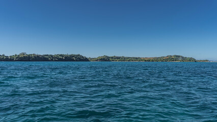 Obraz premium An island on the horizon. Sandy beach. The hills are covered with green tropical vegetation. Clear azure sky. The blue ocean is in the foreground. Ripples on the surface of the water. Madagascar.