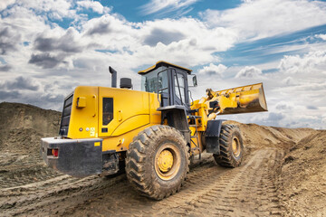 A yellow loader works on a construction site, moving earth and building materials, with clouds in the sky and dirt surrounding the area