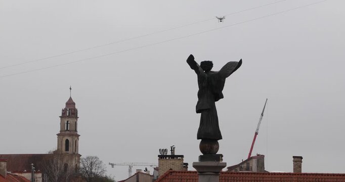 Low-angle view of a bronze angel blowing a horn while a drone films the statue against a flat gray sky. Old meets new&mdash;sacred art and modern surveillance&mdash;evoking tension, wonder, and a futuristic omen.