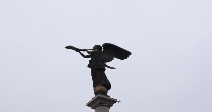 Low-angle view of a bronze angel blowing a horn while a drone films the statue against a flat gray sky. Old meets new&mdash;sacred art and modern surveillance&mdash;evoking tension, wonder, and a futuristic omen.