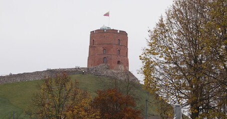 Gediminas Tower stands on its grassy hill in Vilnius as the Lithuanian flag flies in a gray autumn sky. Rustic brick, fortress walls and yellow leaves frame a calm, historic city landmark.