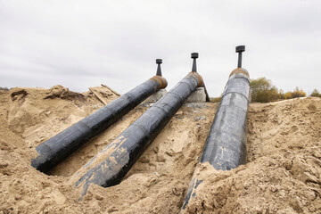 Three large black pipelines lie on a sandy surface at a construction site. The cloudy sky indicates...