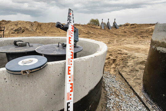 A construction site displays a concrete drainage structure with a measuring stick nearby and sand piled up in the background under a cloudy sky