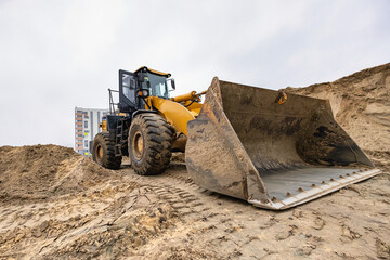 A large loader works at a construction site, moving soil with a bucket in front of a building in an urban area on a cloudy day.