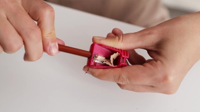 A person sharpens a brown pencil with a red sharpener at a clean desk. This activity showcases attention to detail for art or writing tasks during the day