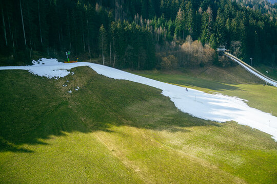 Snow remains on the ski slope as green grass emerges in a mountainous area during spring. Tall trees surround the scene, and a ski lift is visible in the distance