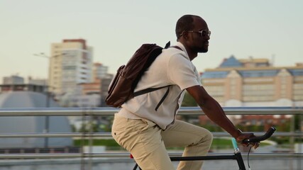 Black businessman with sunglasses and backpack cycling along urban street heading to work on summer day