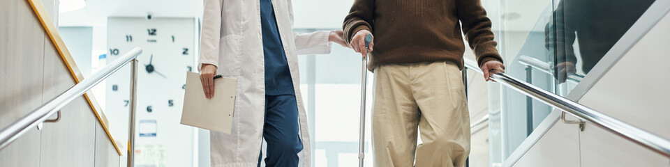 Header of Senior Caucasian man using walking cane assisted by young female doctor walking down...