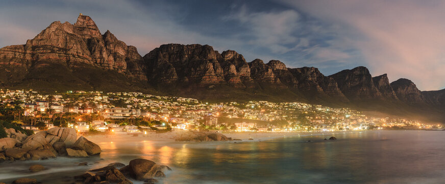 View of the city lights twinkling like scattered jewels beneath the imposing silhouette of a rugged mountain range, reflected in the tranquil waters, Cape Town, Western Cape, South Africa.
