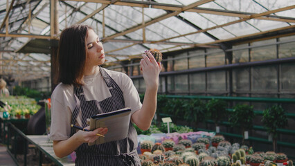 Woman examines cactus while taking notes in a greenhouse filled with various plants during daylight in a vibrant botanical setting