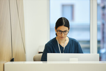 Caucasian young female nurse wearing glasses working on laptop at reception desk in medical office at clinic, focused on screen, dark hair tied back, professional environment visible