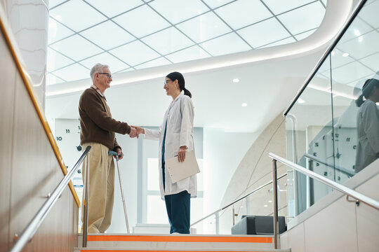 Senior Caucasian man using cane shaking hands with young adult Asian woman doctor standing in modern medical facility, both smiling and making eye contact on staircase, after medical appointment