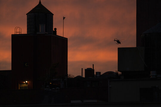 View of a helicopter flying past rooftops and water towers against a vibrant orange and red sky at dusk, New York, New York, United States.