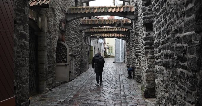 Historic European alley framed by stacked roofed arches. Wet cobblestones lead the eye through textured stone walls as two pedestrians pass, creating depth, symmetry, and a moody overcast atmosphere.