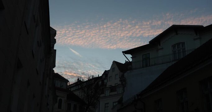 Twilight over an old European street: pink altocumulus clouds and a faint contrail glow above dark rooflines and balconies, forming a serene blue-hour city silhouette with a tranquil mood.