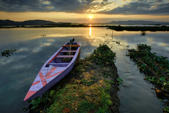 View of a serene lake reflecting the sunset sky, with a small boat gently resting near a grassy bank in the tranquil scene, Ambarawa, Central Java, Indonesia.